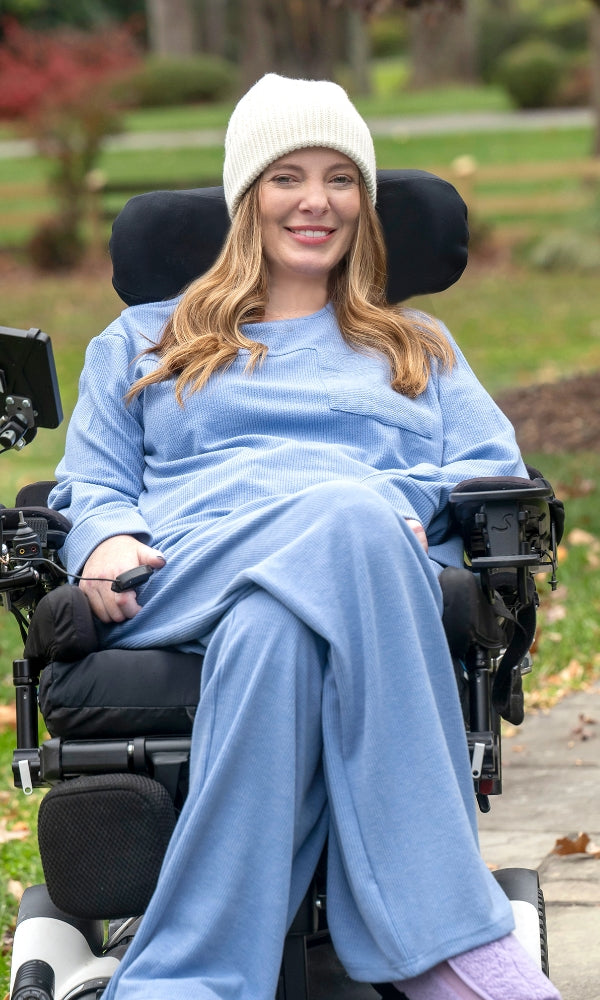 Woman in a wheelchair wearing a light blue outfit and white knit hat outdoors.