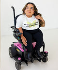 A woman with long brown hair sits in a pink and black electric wheelchair, wearing a white graphic t-shirt that reads 'MY NORMAL IS NOT YOUR NORMAL' with a yellow heart design, paired with black leggings and black boots.