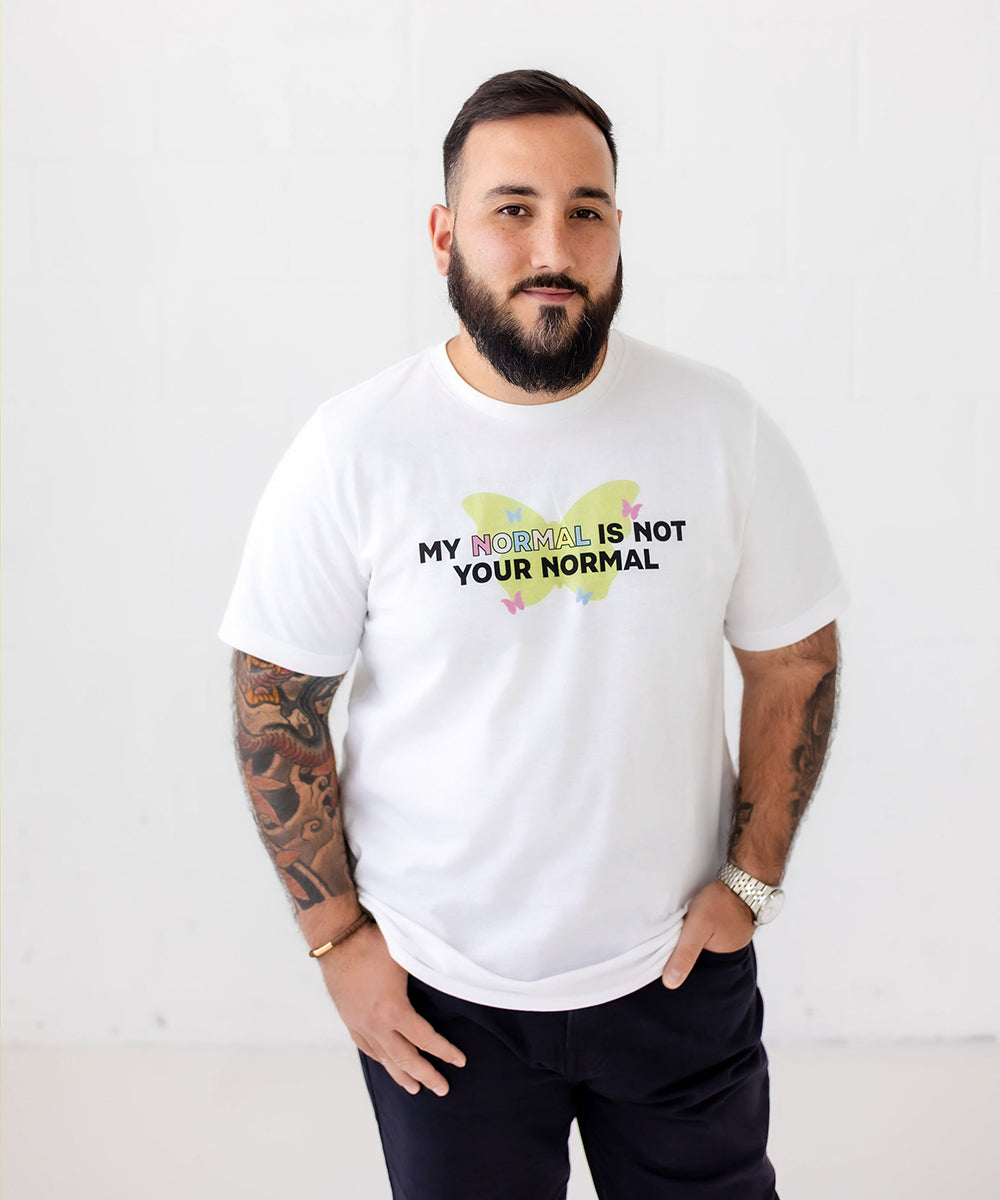 A man with a beard and tattoos wearing a white graphic t-shirt that reads 'MY MOTHER IS NOT YOUR NORMAL' in black text over a yellow and pink heart design, standing against a white wall.