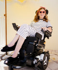 A woman wearing a black and white striped dress, black slide sandals, and a pink headband sits in a black motorized wheelchair. She is smiling and posing on a tiled patio against a pale yellow wall.