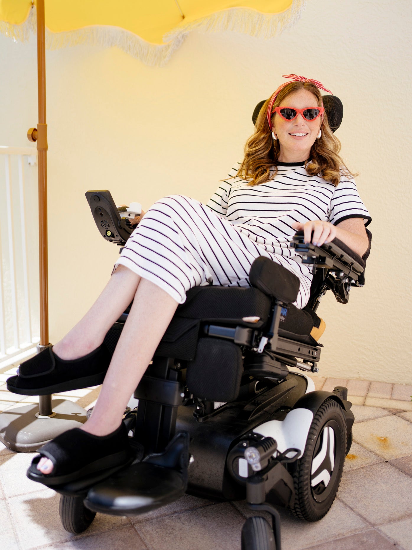 A woman wearing a black and white striped dress, black slide sandals, and a pink headband sits in a black motorized wheelchair. She is smiling and posing on a tiled patio against a pale yellow wall.