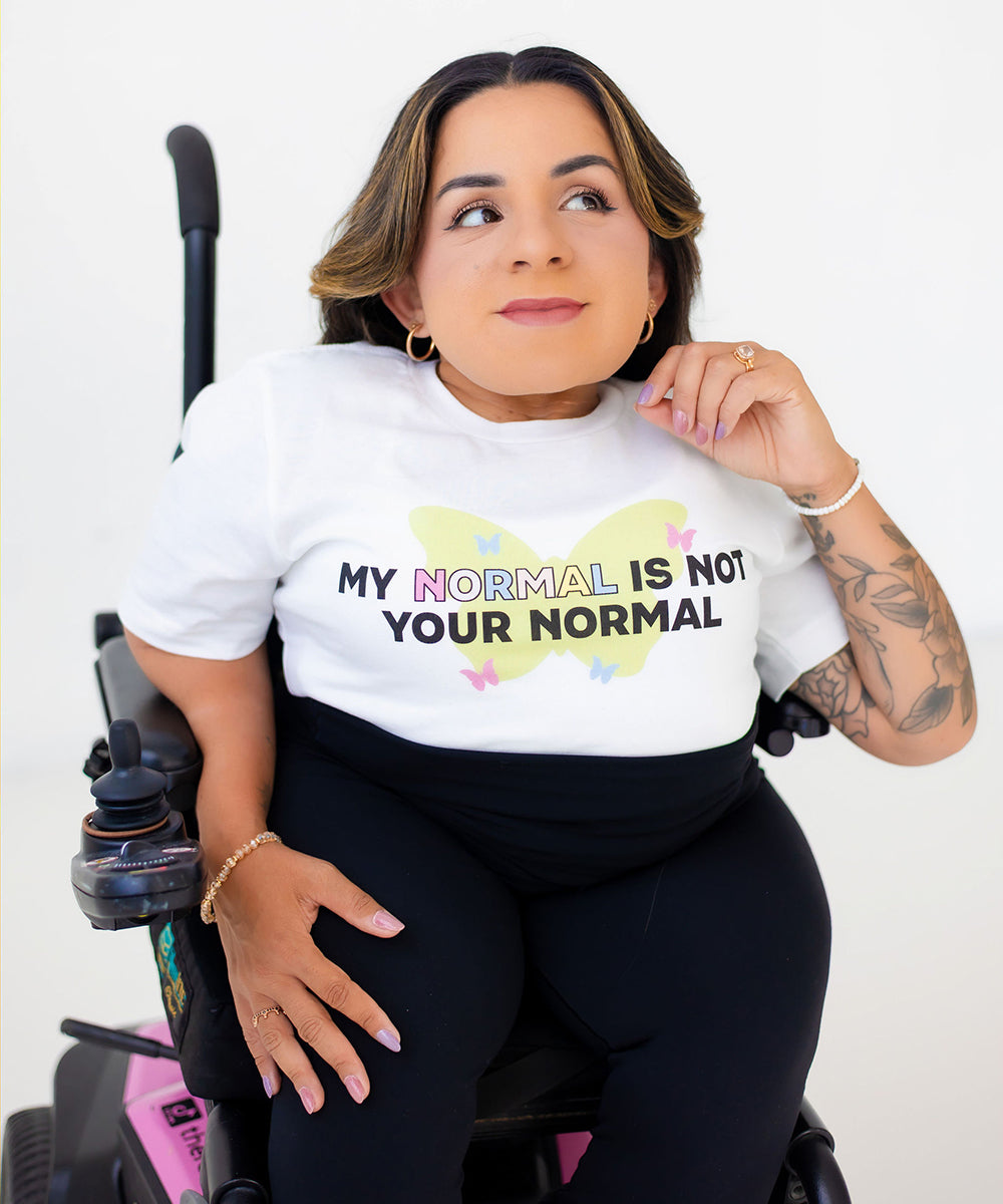 A woman in a wheelchair wearing a white graphic t-shirt with the text 'MY NORMAL IS NOT YOUR NORMAL' printed over a pastel butterfly design. She is wearing black leggings and sitting in a black and pink wheelchair against a plain white background.
