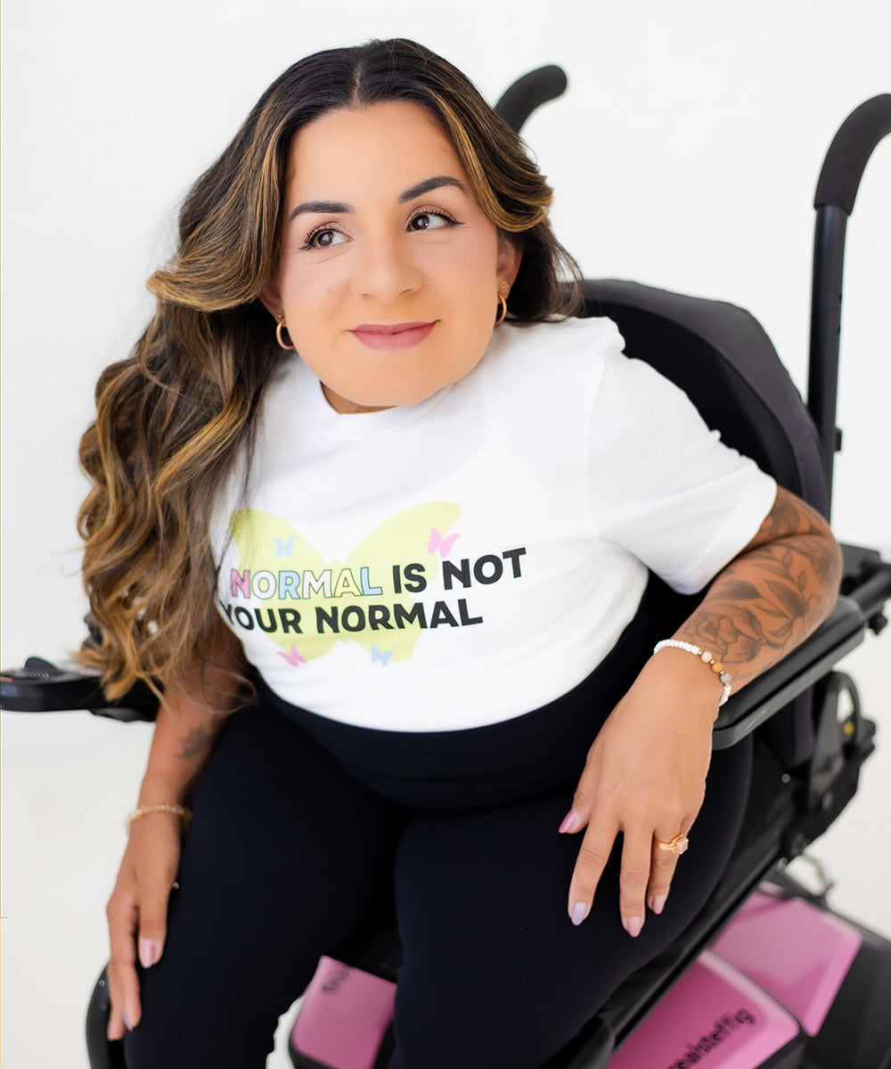 A woman with long wavy brown hair sits in a black motorized wheelchair, wearing a white graphic t-shirt that reads 'NORMAL IS NOT YOUR NORMAL' with a pastel butterfly design. The background is a clean white studio setting.