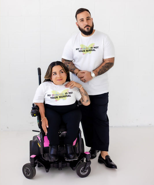 A man and a woman in a motorized wheelchair pose together in a bright white studio. Both are wearing matching white graphic T-shirts with the text 'MY MOTHER IS NOT YOUR NORMAL' and a green and yellow ribbon design.